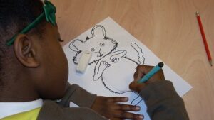 A view over the shoulder of a little girl drawing a mouse character with black pen