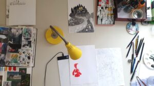 Emily Fellah's desk with paints, pencils, sketchbook, a scene drawn in charcoal and the beginnings of a character drawing