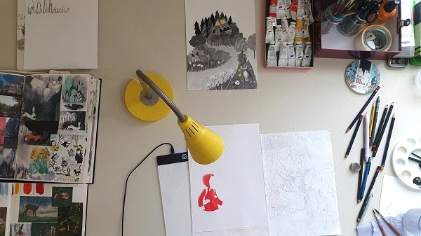 Emily Fellah's desk with paints, pencils, sketchbook, a scene drawn in charcoal and the beginnings of a character drawing
